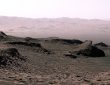 A rover traversing the red rocky surface of Mars with layered terrain and a dusty horizon behind it.