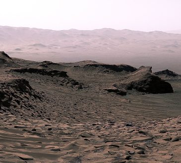 A rover traversing the red rocky surface of Mars with layered terrain and a dusty horizon behind it.