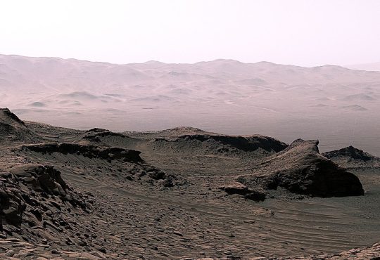 A rover traversing the red rocky surface of Mars with layered terrain and a dusty horizon behind it.